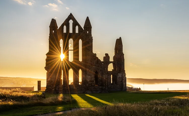 The Gothic ruins of Whitby Abbey with the suns rays producing a star. Sea mist held back by the cliffs at Sandsend in the background