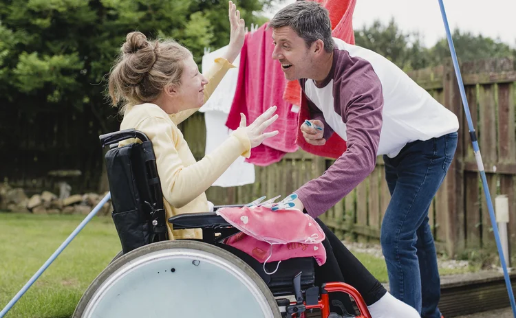 Disabled teenager is in her wheelchair in the garden while her father hangs clothes on the washing line. They are interacting playfully.